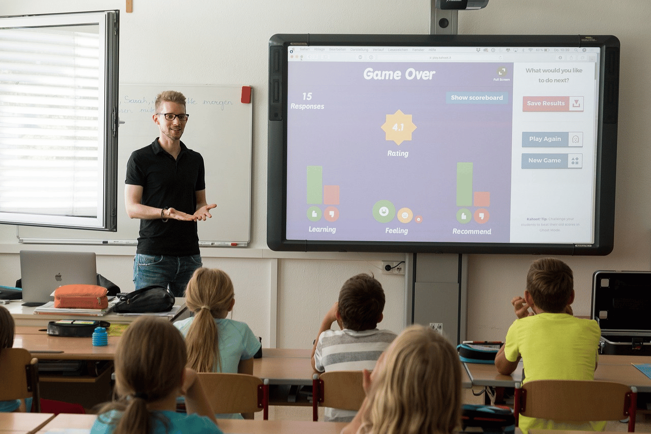 teacher standing in front of the class , learners sitting inside the classroom , big device on the wall